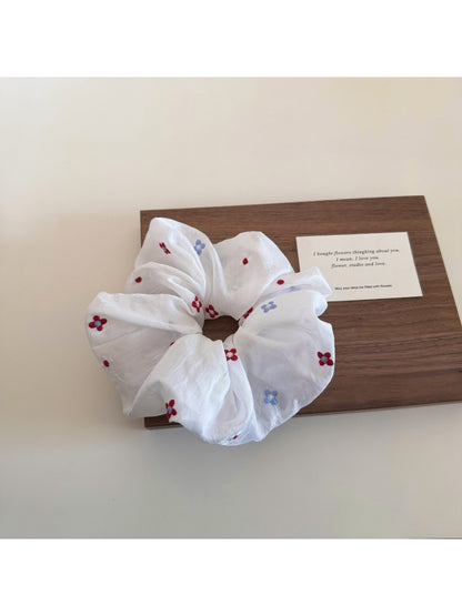 Minimalist white flower scrunchie next to green leaves on a white surface
